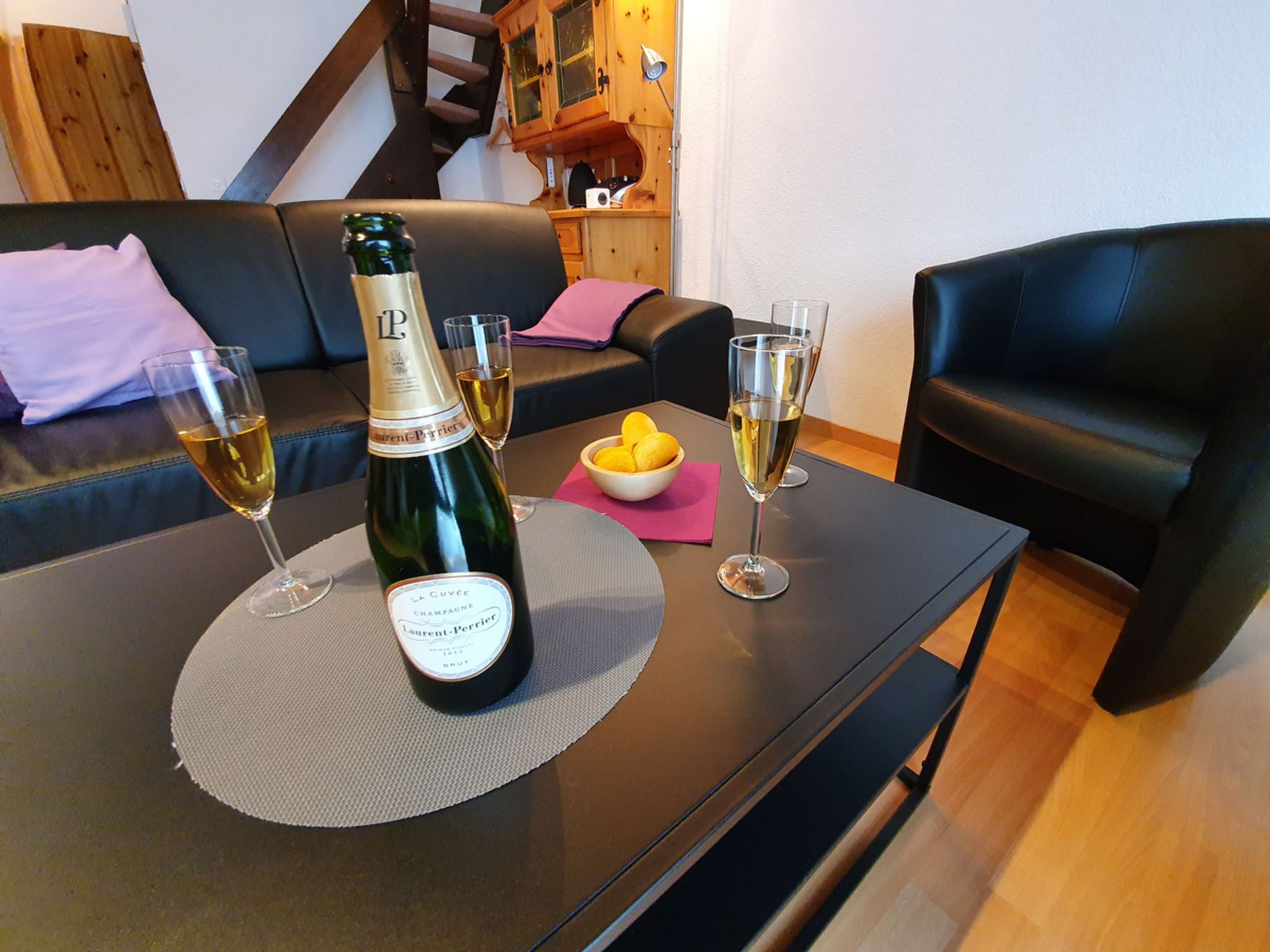 Une bouteille de champagne La Cuvée est posée sur une table, accompagnée de deux verres à champagne, d’un bol de citrons et d’une serviette rose.