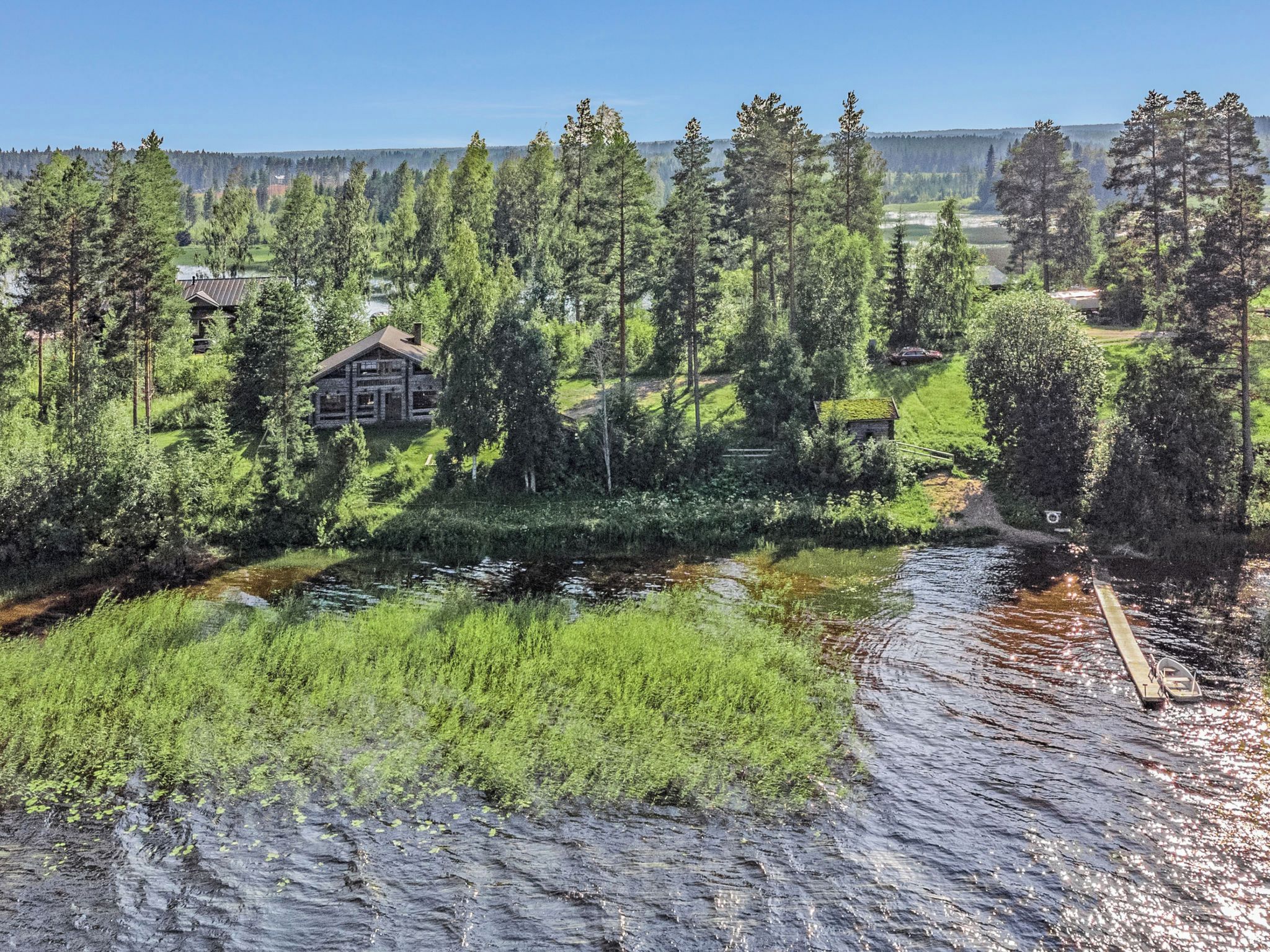 En stuga vid sjön med en brygga, omgiven av tät skog och en klar himmel.