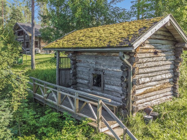 A rustic log cabin with a moss-covered roof, a small window, and a wooden deck leading to the entrance.