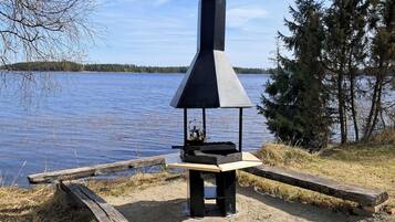 A portable grill with a chimney, set on a gravel area by a lake with trees in the background.