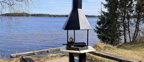 A portable grill with a chimney, set on a gravel area by a lake with trees in the background.