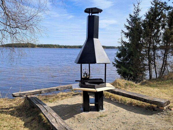 En portabel grill med en skorsten, placerad på ett grusunderlag vid en sjö, med träd i bakgrunden.