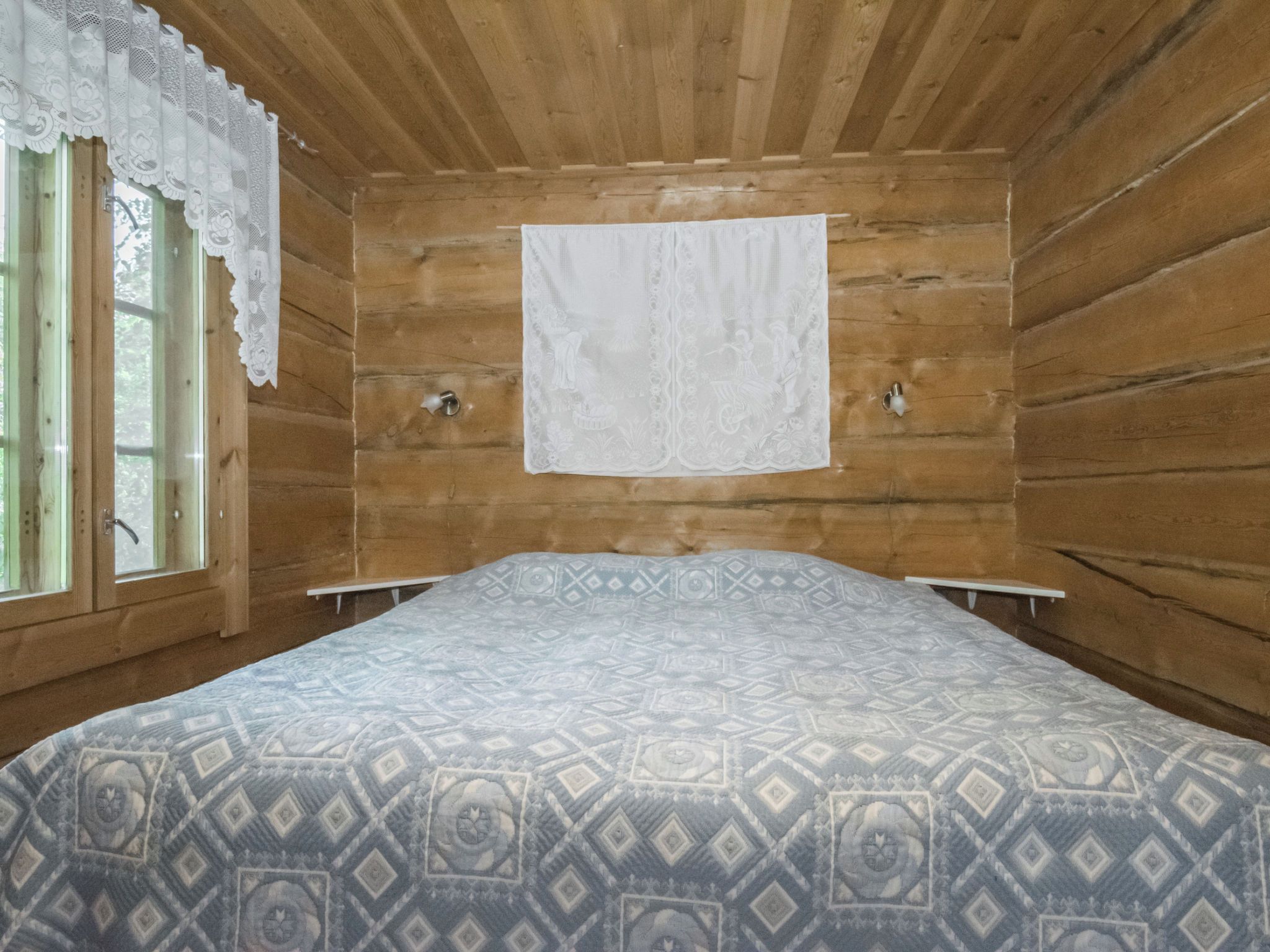 A wooden cabin room with a bed, a window with lace curtains, and a white curtain on the wall.
