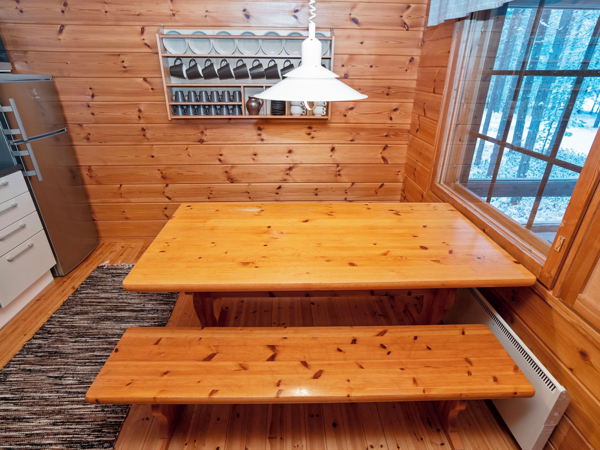A wooden interior with a table and bench, a rack of mugs, and a window with a view of trees.