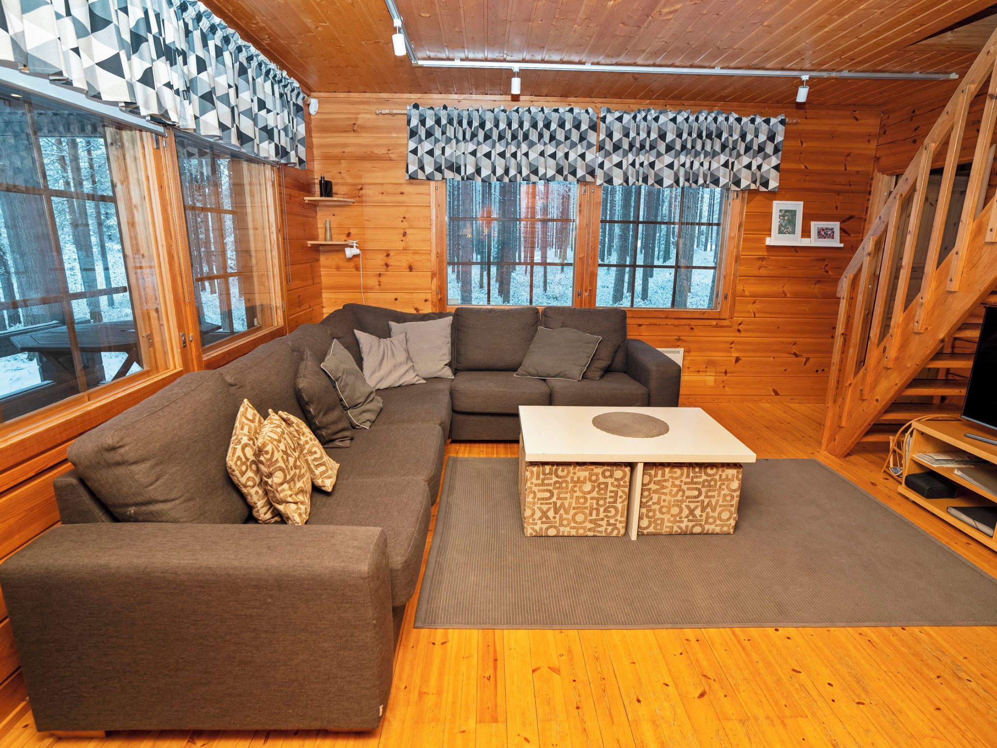 Interior acogedor de una cabaña con un sofá seccional, una mesa de centro y ventanas amplias que ofrecen vistas a un bosque nevado.