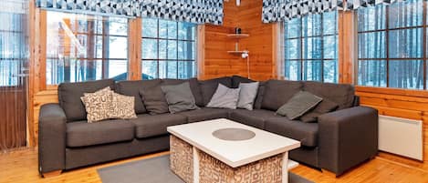 Ein modernes Blockhaus-Interieur mit einer Eckcouch, einem zentralen Couchtisch und großen Fenstern mit geometrischen Vorhängen.