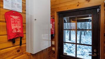 Een houten cabinestijl interieur met een witte koelkast, een brandblusser en een deur die uitkomt op uitzicht over een besneeuwd bos.