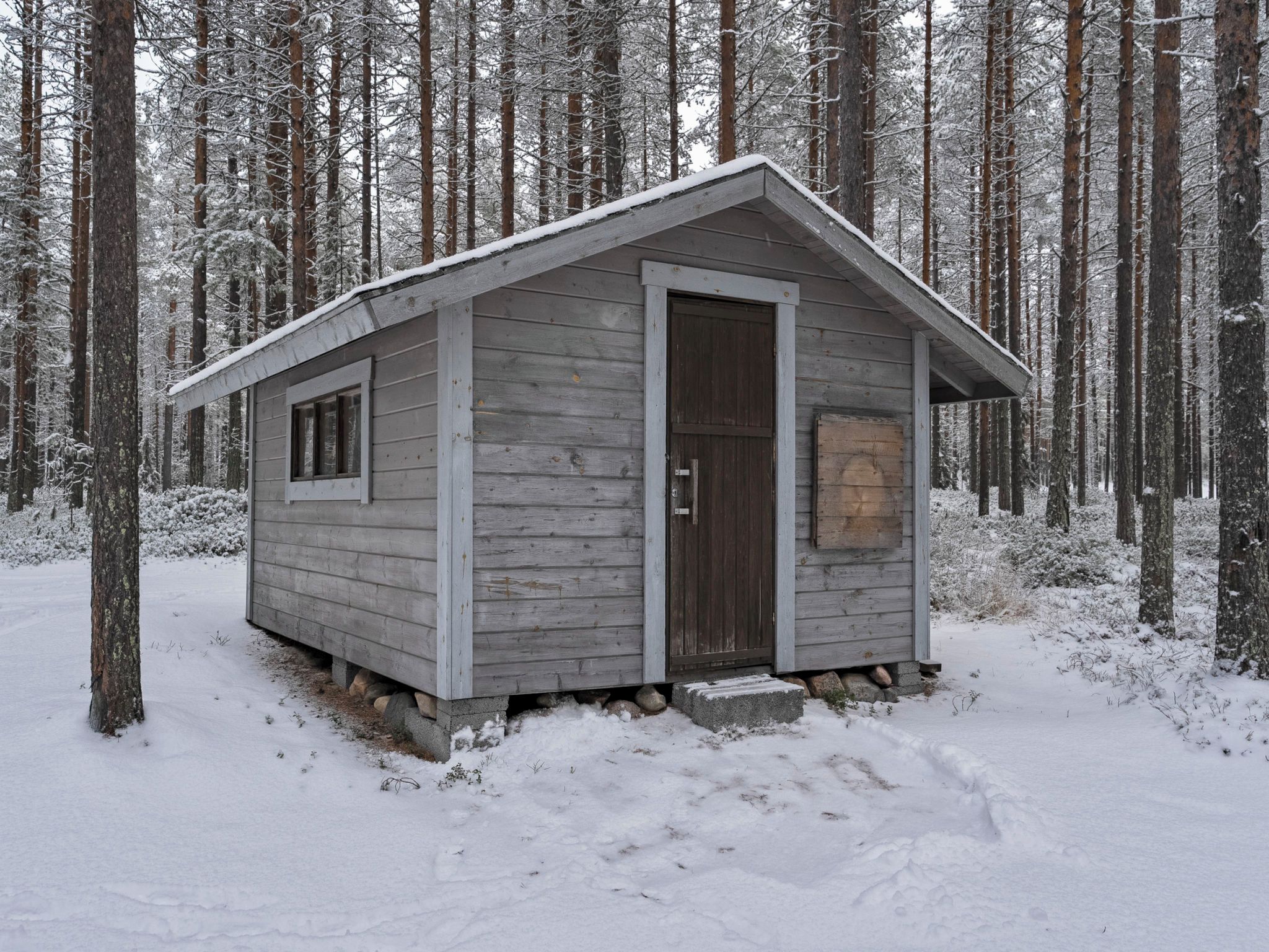 A small wooden cabin with a door and a window, surrounded by snow-covered trees.