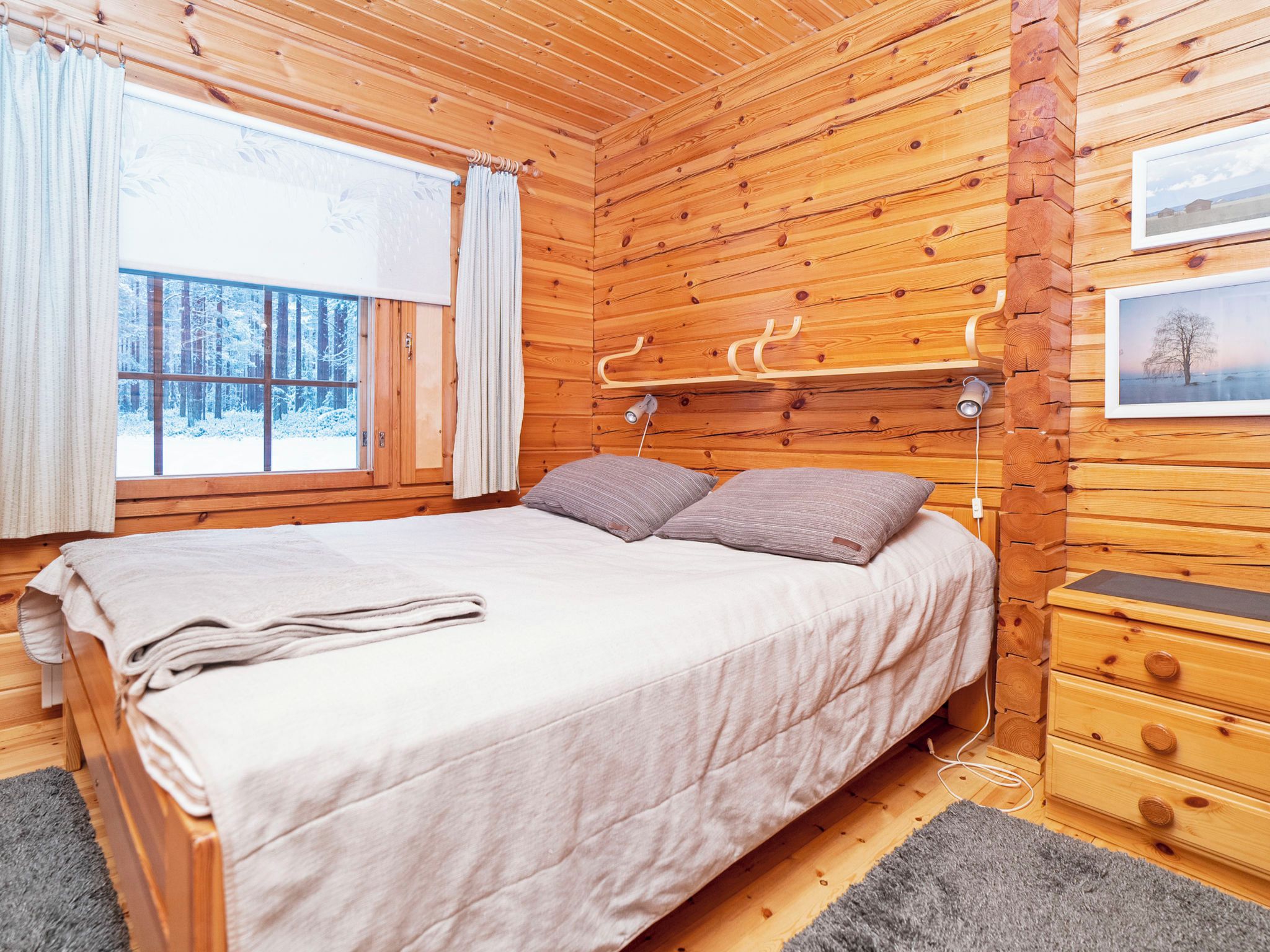 Een slaapkamer in een houten blokhut met een bed, nachtkastje, lamp en een raam met gordijnen.