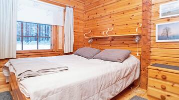Een slaapkamer in een houten blokhut met een bed, nachtkastje, lamp en een raam met gordijnen.