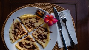 A plate of crepes with chocolate sauce, a knife, a fork, and a napkin on a woven placemat. - Hotra Hill Cottage (Penida Island)
