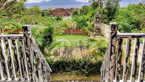 A view from a wooden deck overlooking a lush garden with a house and a mountain in the background. - Hotra Hill Cottage (Penida Island)