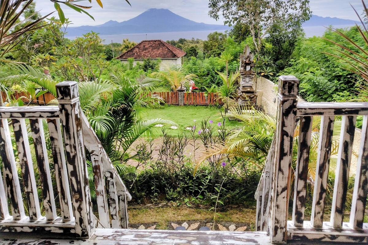 A view from a wooden deck overlooking a lush garden with a house and a mountain in the background.