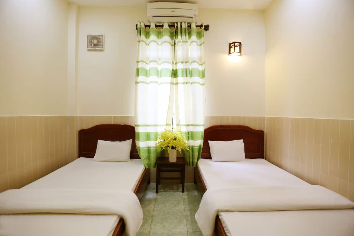 Une chambre d’hôtel avec deux lits simples, une petite table sur laquelle est posé un vase de fleurs, un climatiseur fixé au mur et une fenêtre ornée de rideaux verts et blancs.