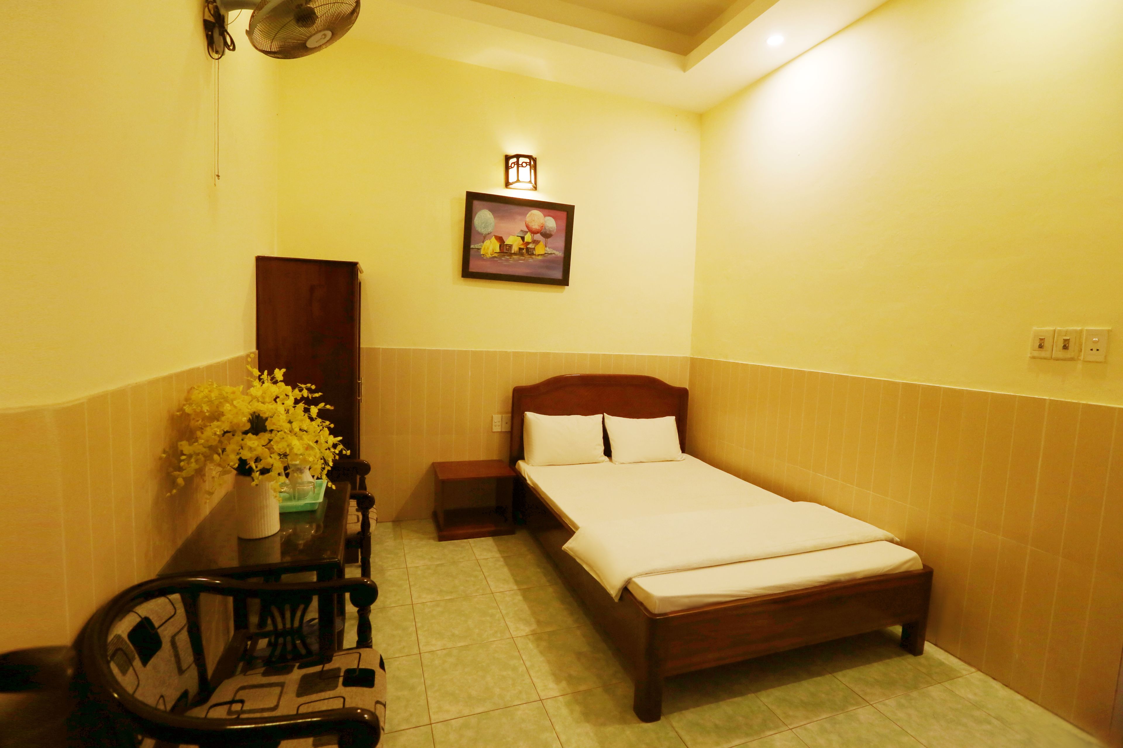 A hotel room with a wooden bed, a nightstand, a chair, a small table with a vase of flowers, and a wall-mounted picture.