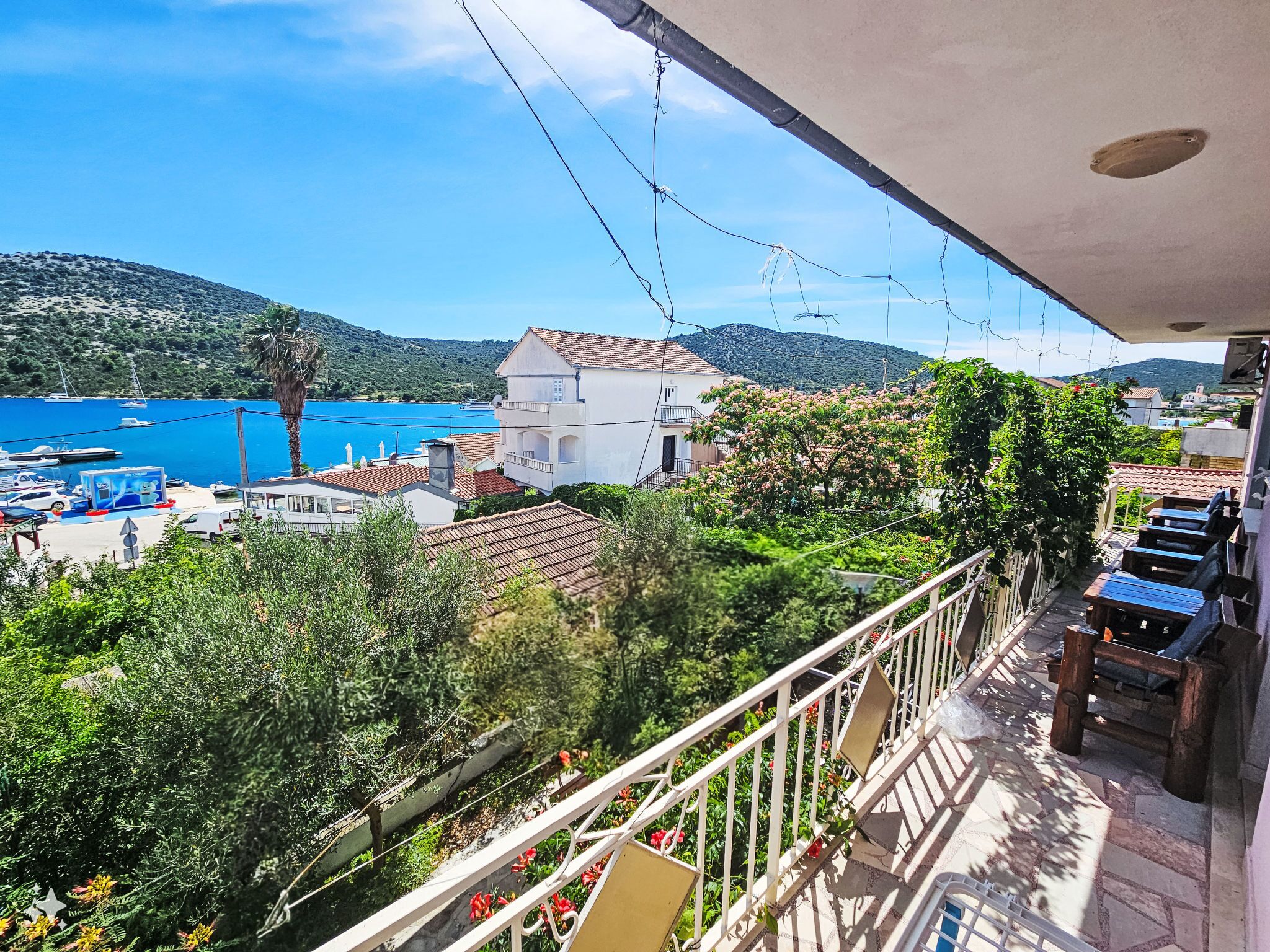 Un balcón con vistas a un pueblo costero, un puerto deportivo con barcos y abundante vegetación.