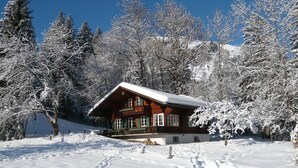 A wooden cabin with a snow-covered roof, surrounded by snow-laden trees and a clear blue sky. - Chalet Maru by Interhome (Lenk)