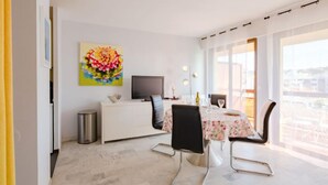 Un comedor moderno con una mesa blanca dispuesta para dos, un televisor sobre un mueble blanco y un amplio ventanal con vista a la ciudad.