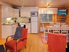 A compact kitchen with white cabinets, a stove, a microwave, and a refrigerator. There is a dining area with a wooden table and chairs, and a television mounted on the wall. - Suopursu by Interhome (Kuusamo)