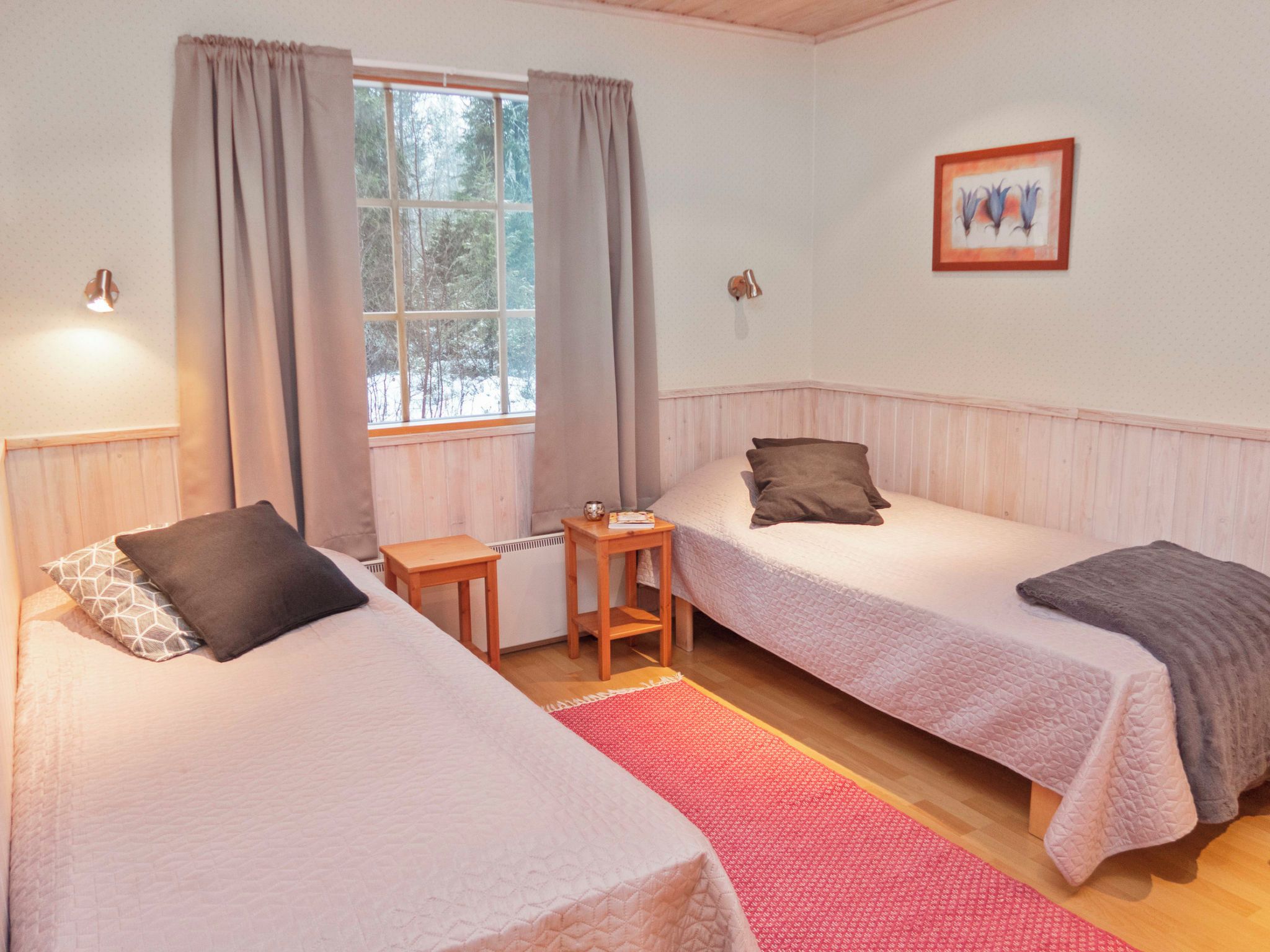 Ein Zimmer mit zwei Betten, einem kleinen Tisch, einem Fenster mit Vorhängen und einem Wandbild.