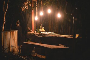 An outdoor dining area with a rustic wooden table set for two, surrounded by trees and illuminated by hanging light bulbs. - Regenta Resort Aranyaani (Kurai)
