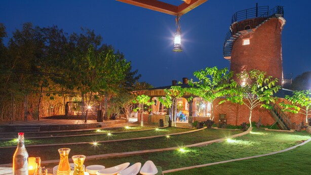 A well-lit outdoor dining area with a rustic brick building, a wooden table set with dinnerware and candles, and a staircase leading to a tower. - Regenta Resort Aranyaani (Kurai)