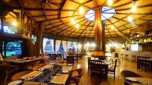 A restaurant with a rustic interior, featuring wooden tables and chairs, a large central light fixture, and a television screen displaying a scenic view. - Regenta Resort Aranyaani (Kurai)