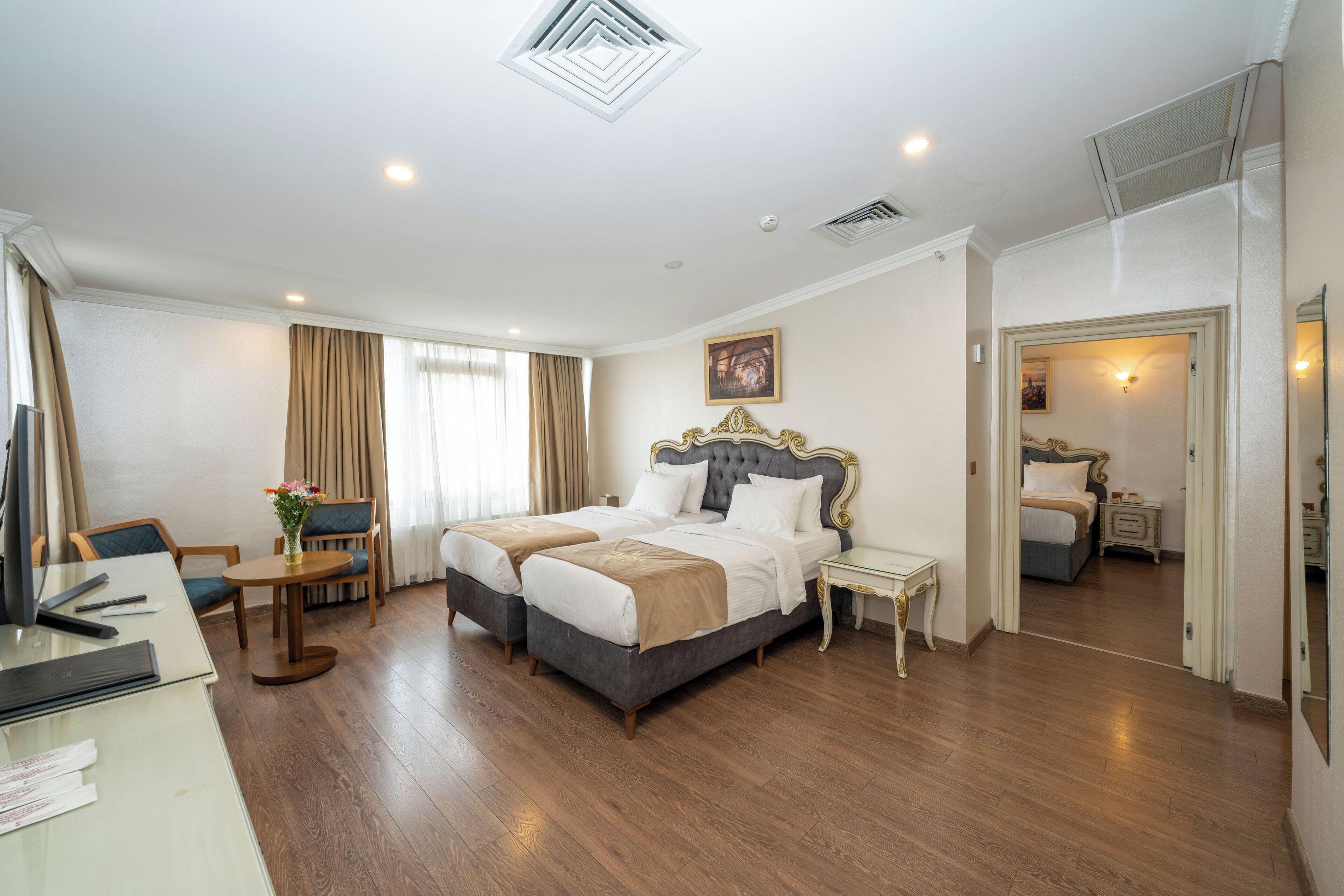 A hotel room with a double bed, a desk with a computer, a chair, a small table with a vase of flowers, and a mirror.