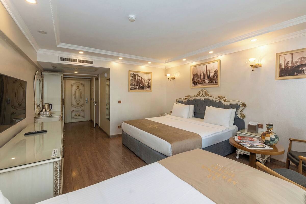 A hotel room with a large bed, a smaller bed, a desk with a chair, a mirror, a television, and a door leading to another room.