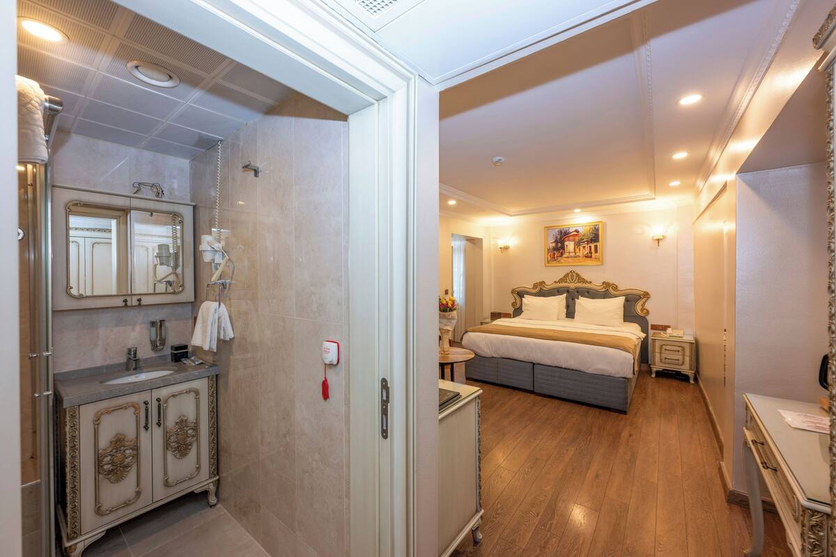 A hotel room with a bathroom featuring a sink, mirror, and vanity. The bedroom has a bed, nightstands, and a painting on the wall.