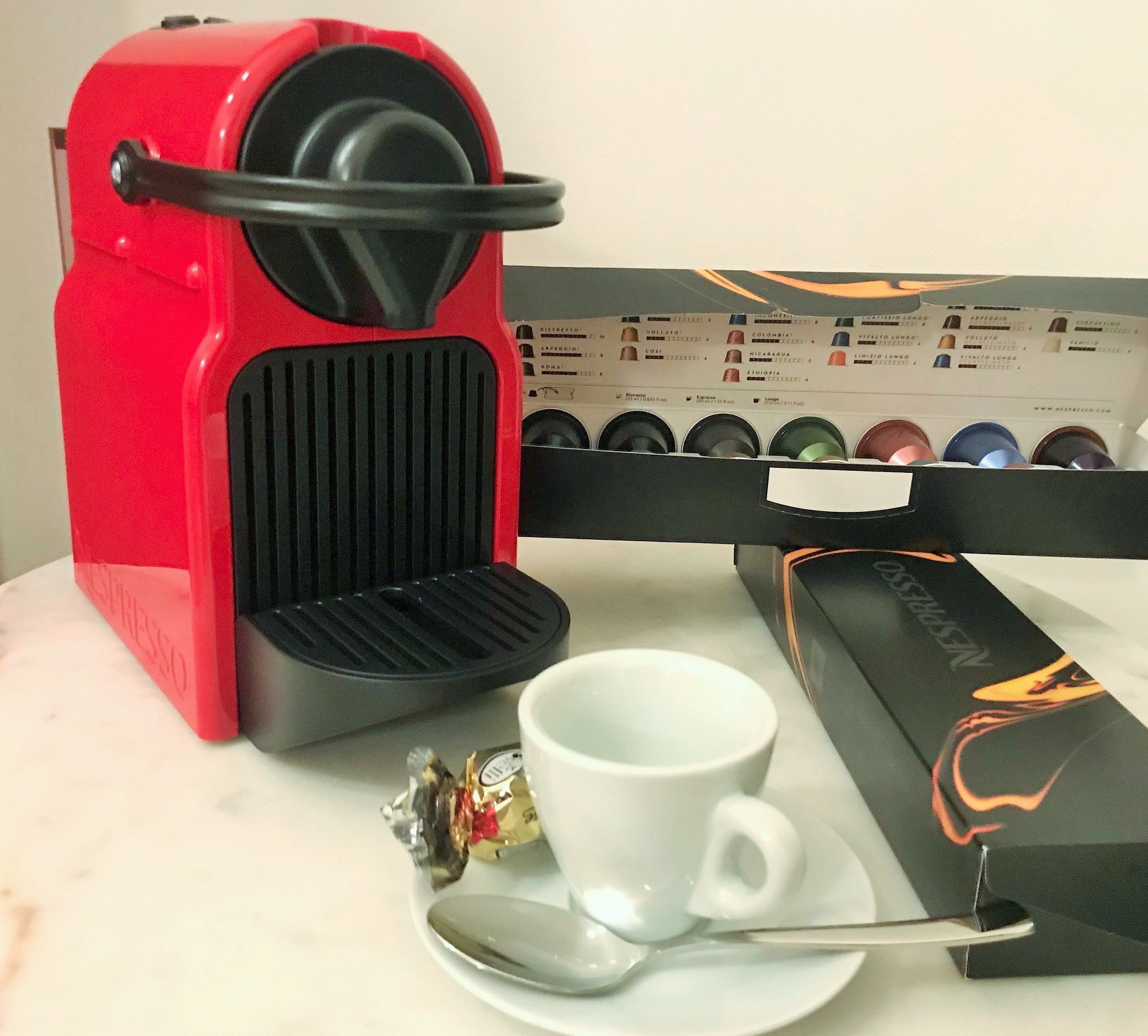 Une machine à expresso rouge, une tasse blanche, une cuillère et une boîte de capsules de café, le tout posé sur une surface en marbre.