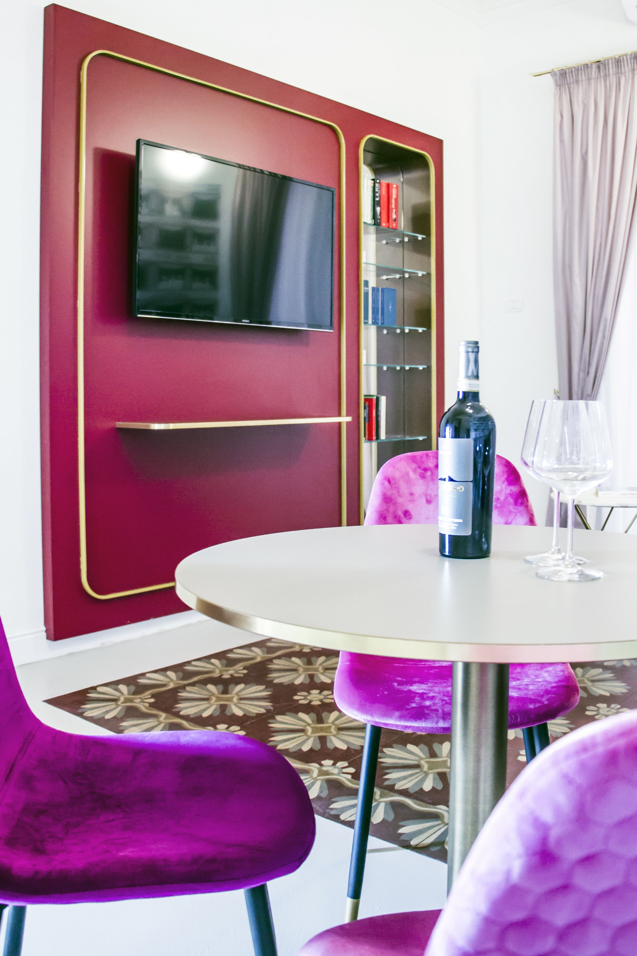 Una stanza con un tavolo rotondo, una bottiglia di vino e un bicchiere da vino. C'è una televisione montata su un muro rosso con decorazioni dorate. La stanza presenta sedie viola e un tappeto decorato.