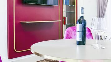 Una stanza con un tavolo rotondo, una bottiglia di vino e un bicchiere da vino. C'è una televisione montata su un muro rosso con decorazioni dorate. La stanza presenta sedie viola e un tappeto decorato.