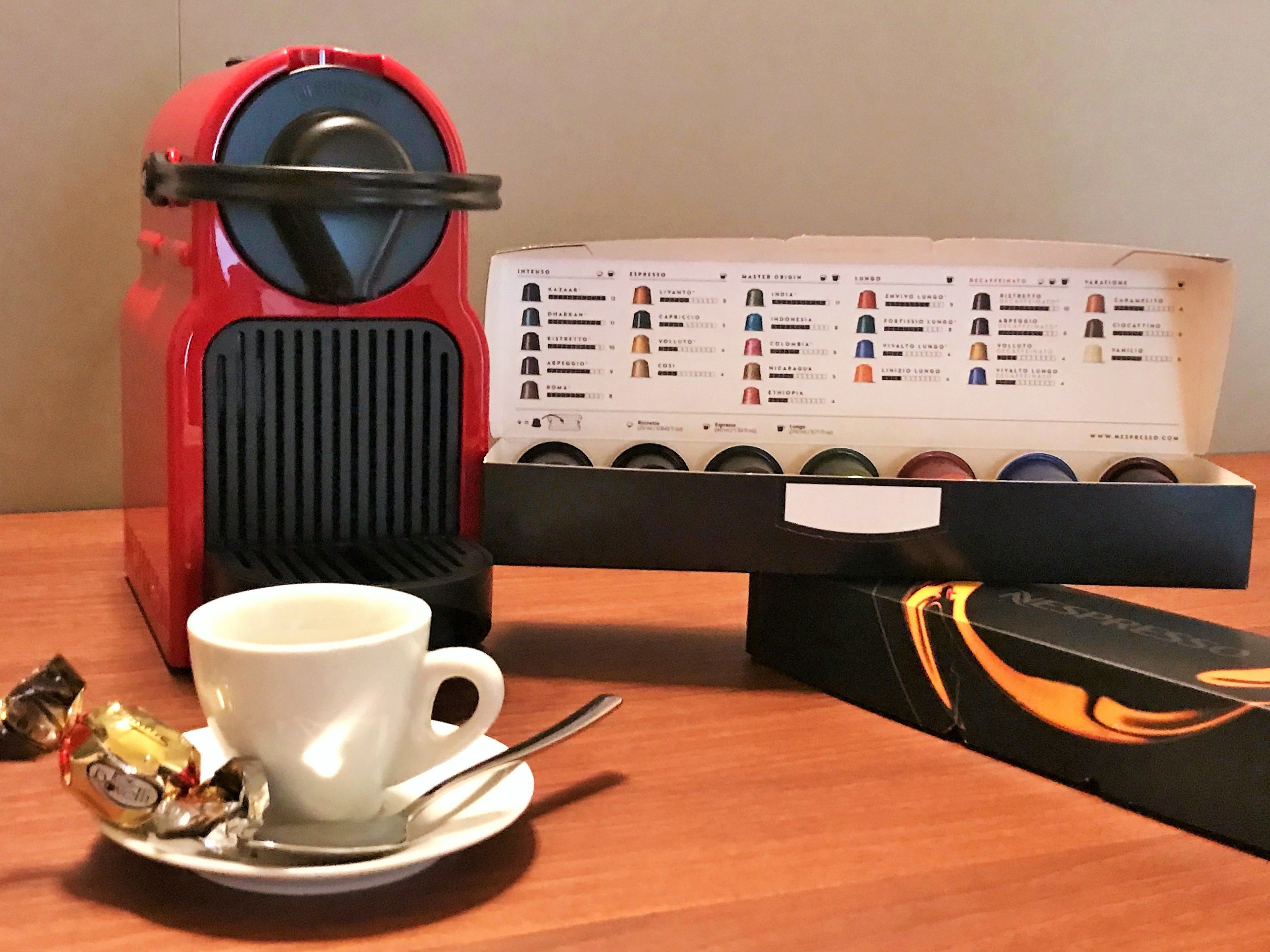 A coffee maker, a coffee pod box, a cup, and a spoon on a wooden table.