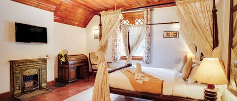 A room with a wooden ceiling, a bed with white bedding, a bedside table with a lamp, a fireplace, and a television on the wall.