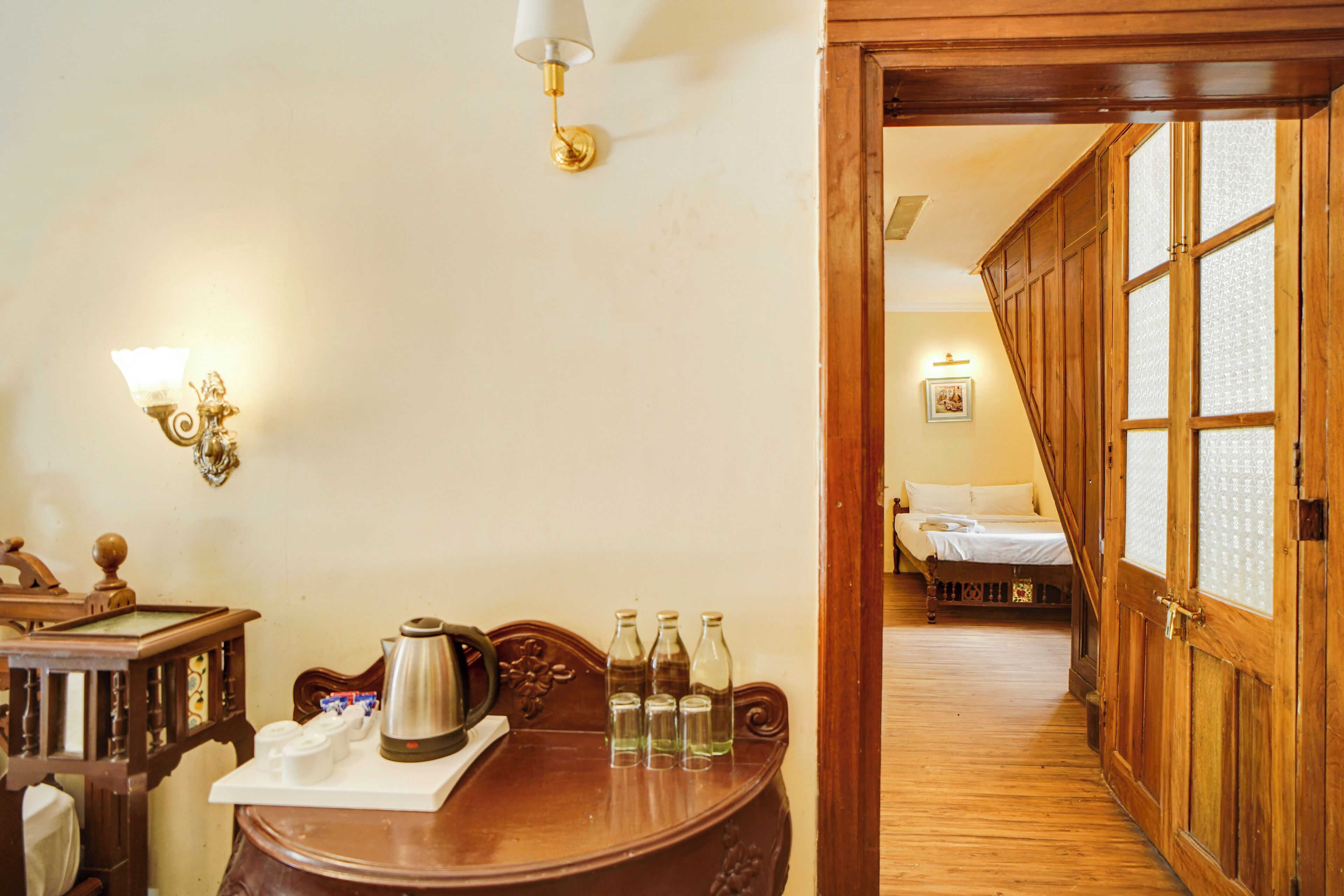 A wooden table with a coffee pot and bottles, a wall-mounted light fixture, and a wooden door leading to a bedroom with a bed and a framed picture.