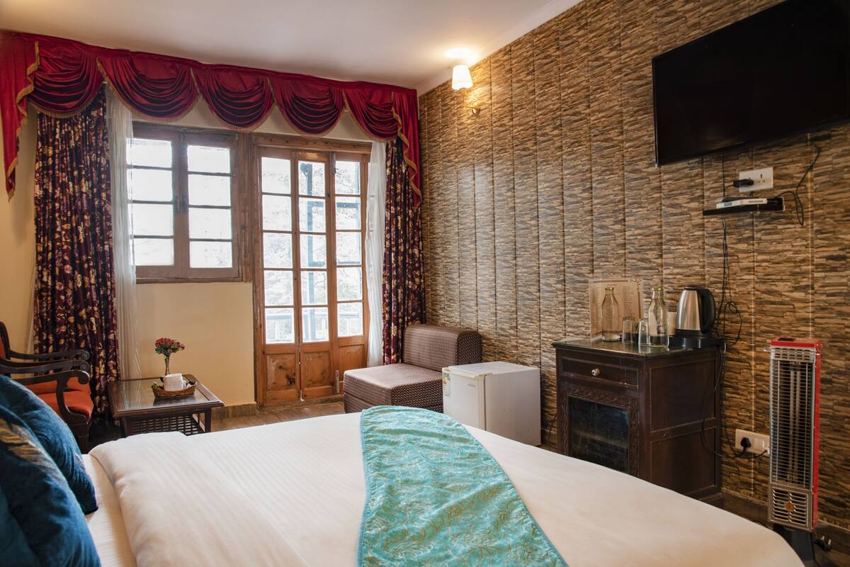 A hotel room with a large bed, a wooden dresser, a small table with a vase, a chair, a window with curtains, and a flat-screen TV mounted on the wall.