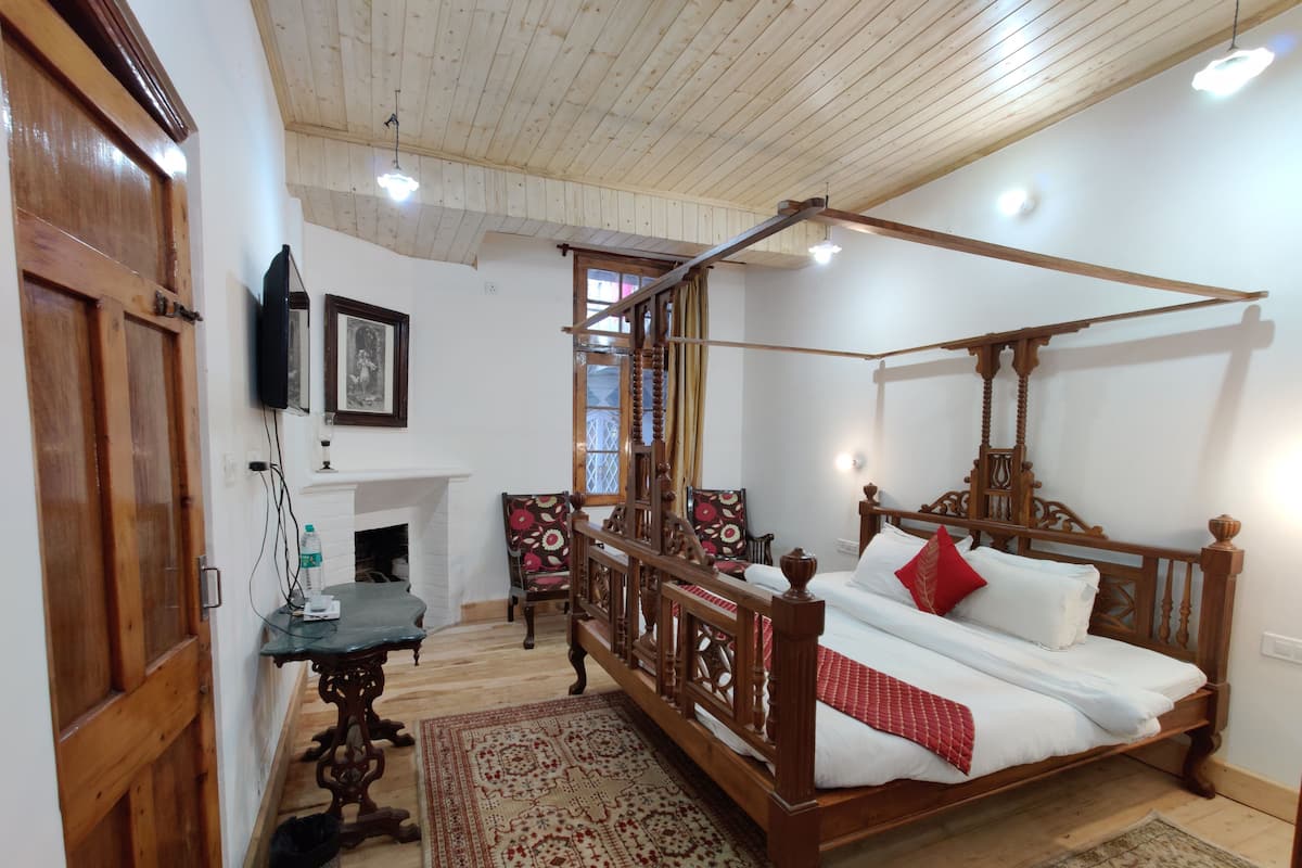 A four-poster bed with a wooden frame, a white mattress, and red pillows. There is a small table with a glass top and a chair with a floral pattern. A fireplace is visible in the background, and a television is mounted on the wall.
