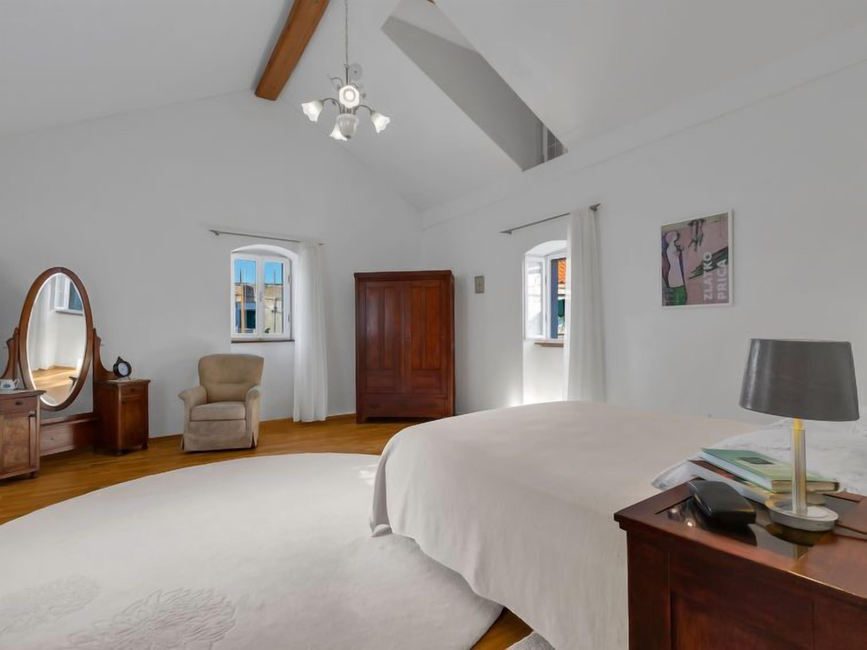 A spacious bedroom with a large bed, a wooden wardrobe, a chair, and a desk with a lamp.