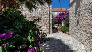 A narrow cobblestone alley flanked by stone walls and lush greenery, leading to a building with a red-tiled roof. - Marijan by Interhome (Brač/Sutivan)