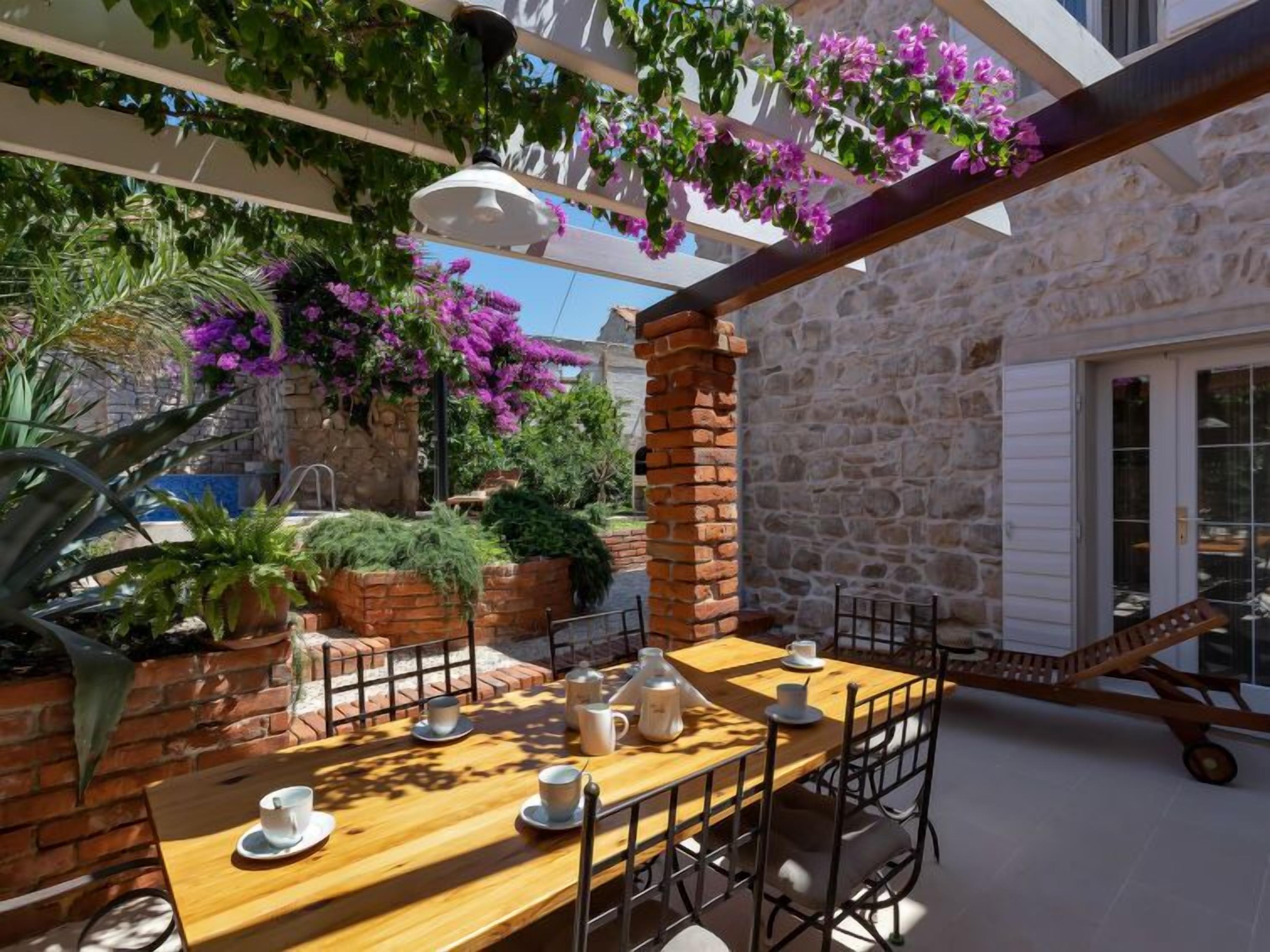 An outdoor patio with a wooden table set for two, surrounded by stone walls and potted plants.