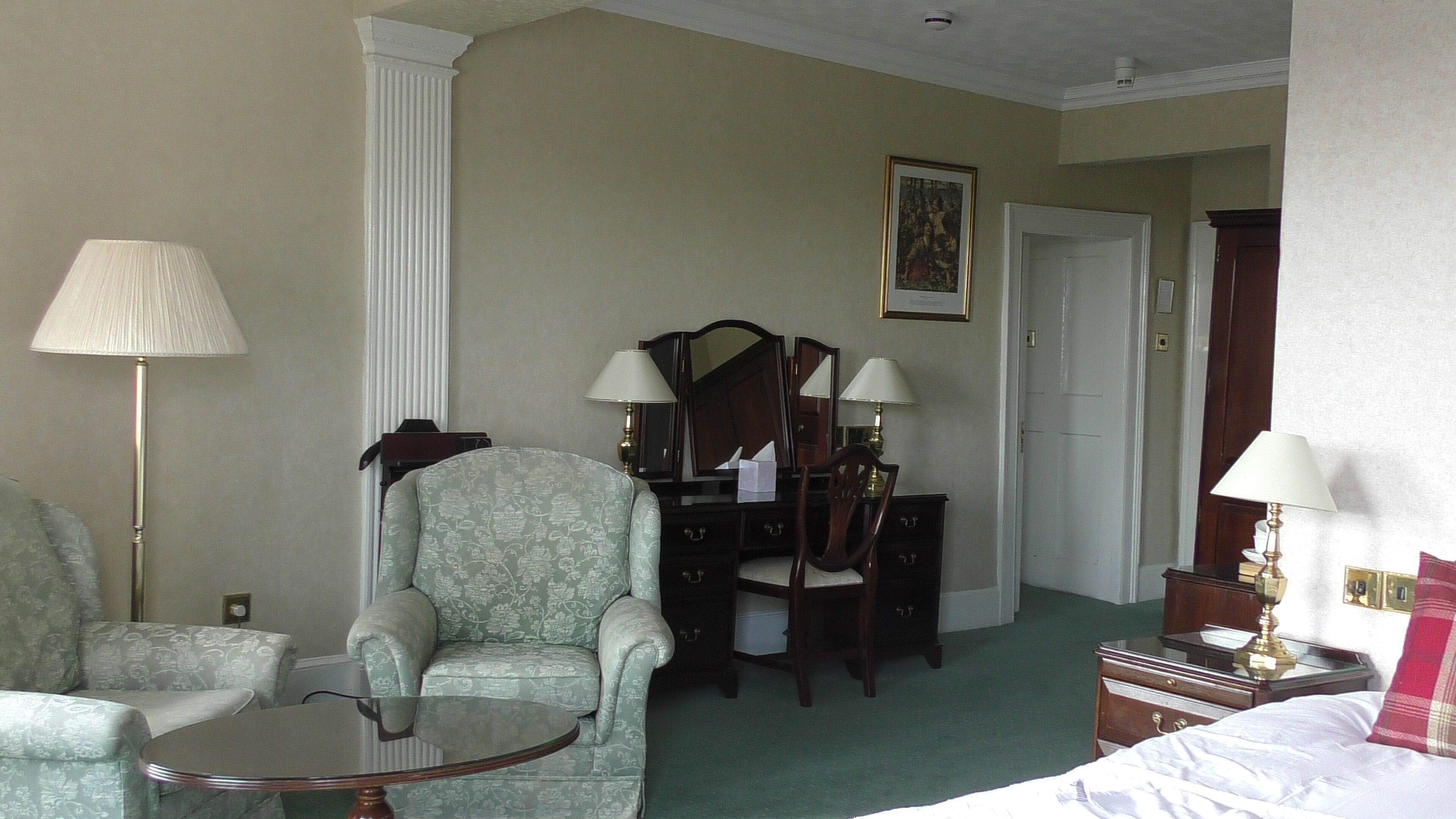 A hotel room with a green patterned armchair, a small round table, a wooden dresser with a mirror, and a bed with white bedding.