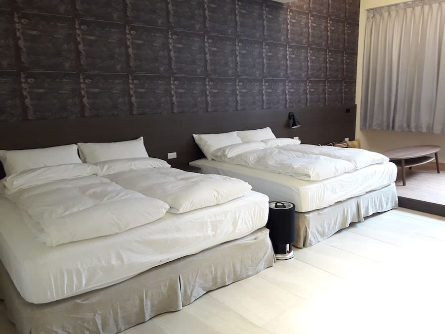 A hotel room with two beds, a dark textured wall, a bedside table, a lamp, and a chair.