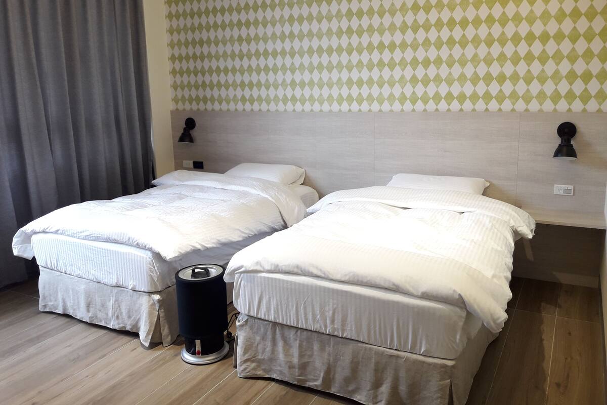 A hotel room with two single beds, a wooden floor, a grey curtain, and a patterned wall.