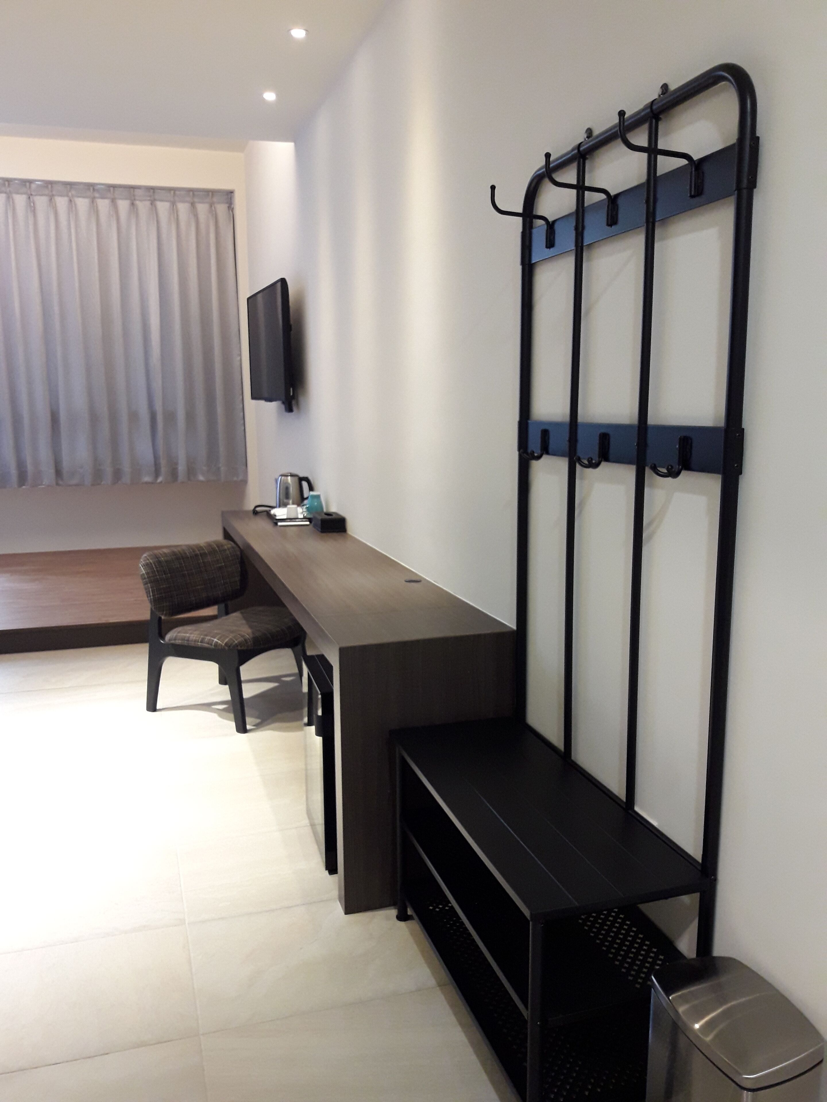 A modern hotel room with a flat-screen TV mounted on the wall, a desk with a chair, a bench, and a trash bin.