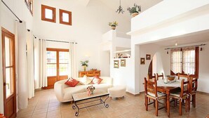 A spacious living room with a sofa, coffee table, and dining table set. There are potted plants, framed pictures, and a chandelier. - Ca Toni by Interhome (Moraira)