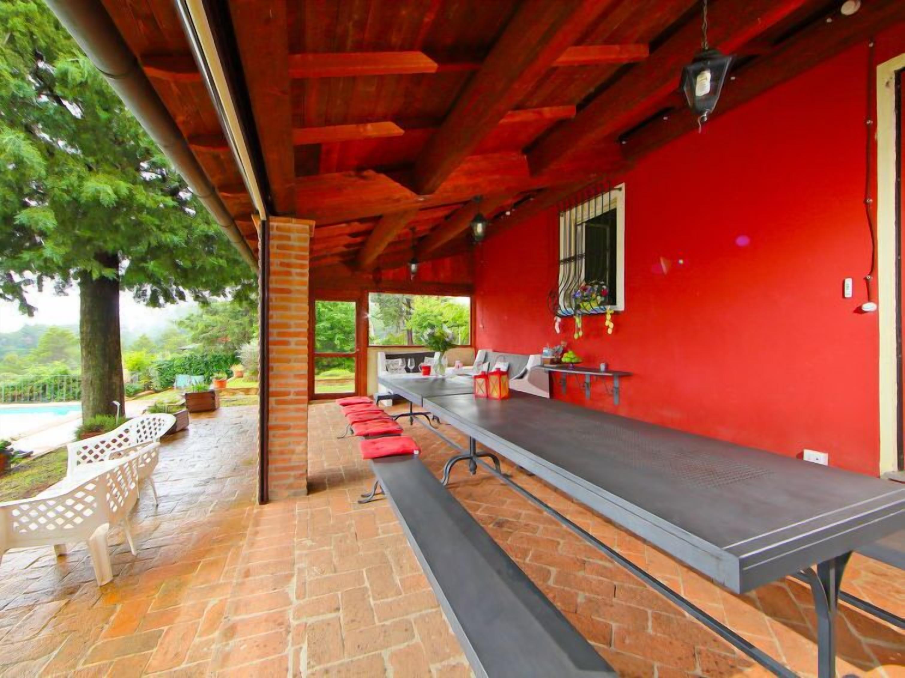Un espace extérieur abrité, aménagé pour les repas, avec une longue table, des murs rouges et une vue sur une piscine.