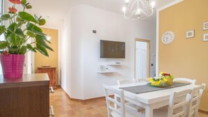 Un coin repas avec une table et des chaises blanches, un téléviseur fixé au mur et une plante dans un pot rose.