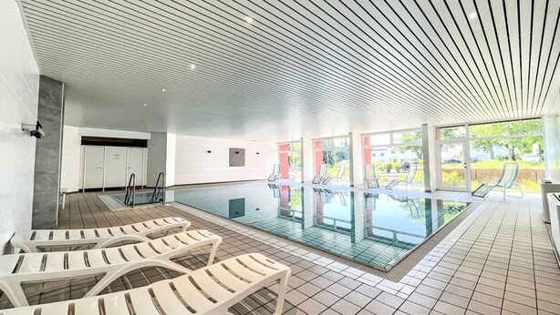Una piscina interior moderna con paredes de vidrio transparente, sillas reclinables blancas y suelo de baldosas.