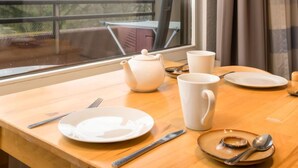 Una mesa de comedor con tetera, tazas, platos, cubiertos y una ventana que ofrece vistas a árboles y un balcón.